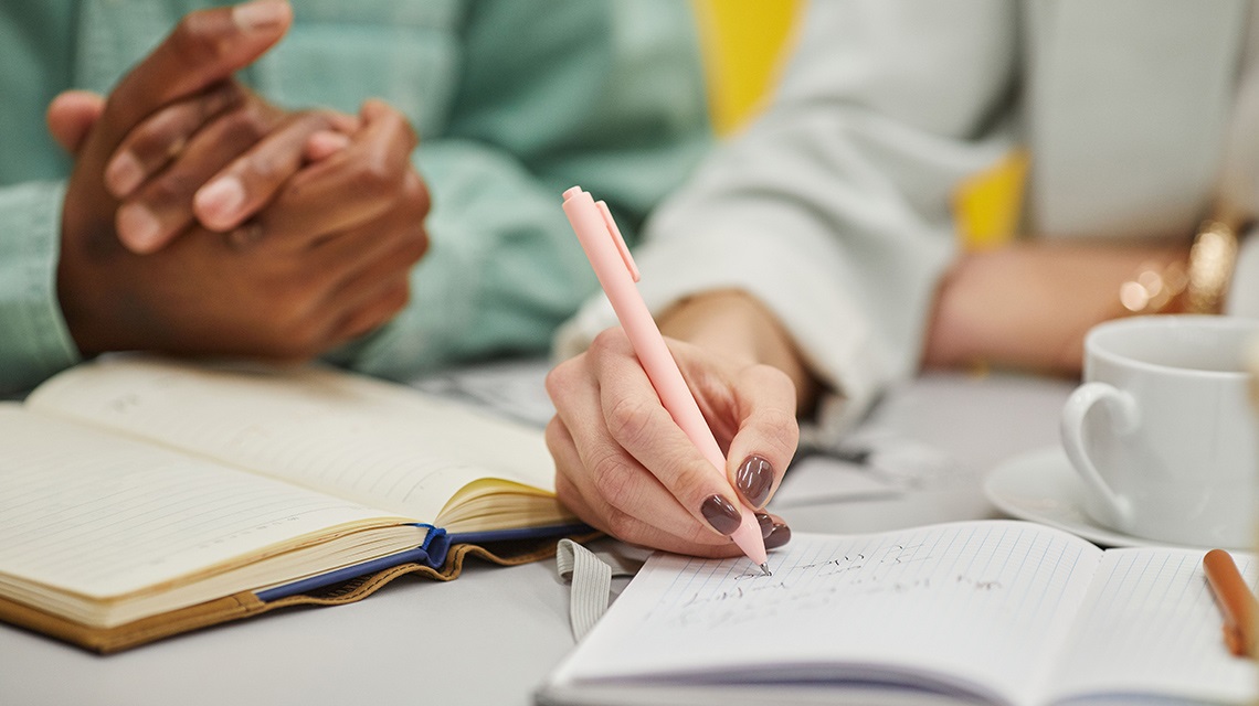 2 people with notebooks in front of them. One of them is writing in the notebook.