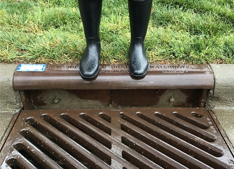 Close-up of a storm drain with someone from the knees down wearing golashes