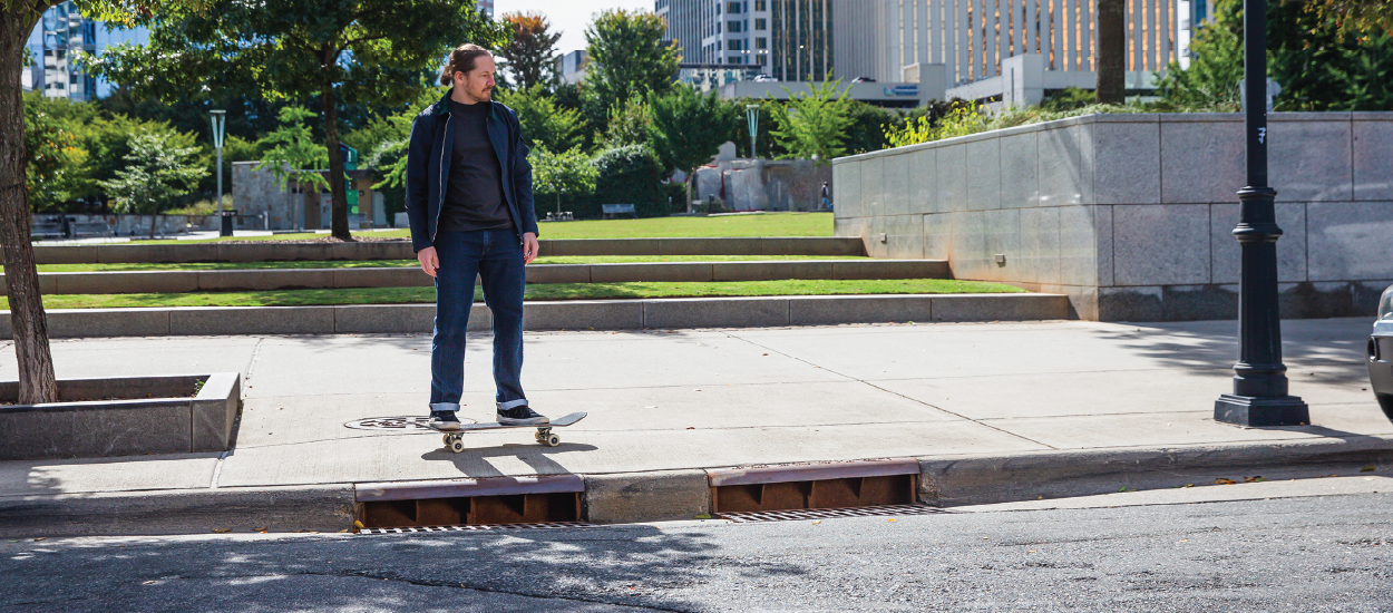 One young man riding a skateboard on the sidewalk with storm drain showing.