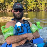a bearded man wearing sunglasses, a baseball cap and inflatable alligator arm cuffs sitting in an innertube on a river