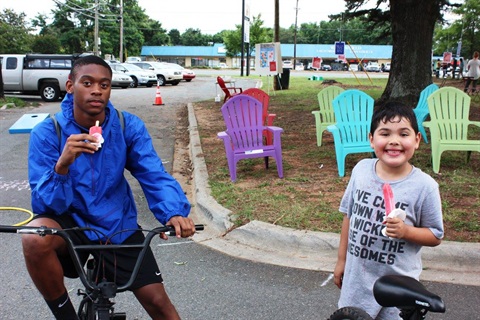 A teenager on his bike and a sweaty kid eating popsicles in front of colorful adironack chairs