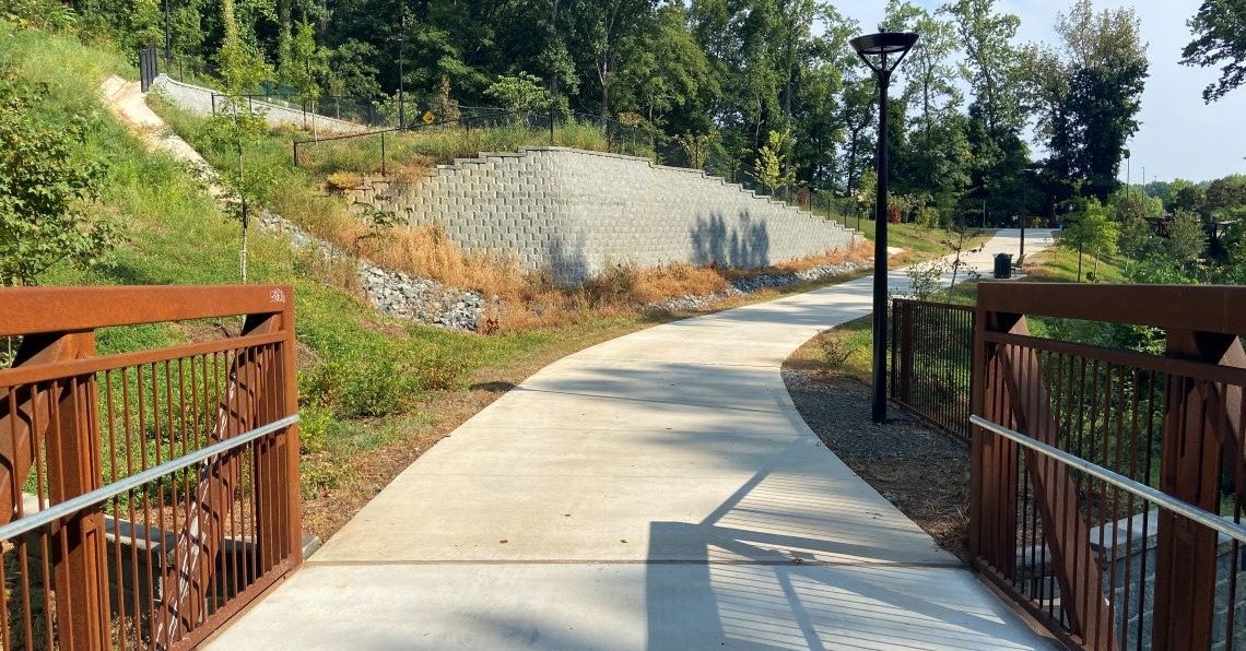 A view of the Oakhurst/Amity Gardens Multiuse Path from a red bridge looking toward the path flanked on the left by a large retaining wall and storm water infrastructure
