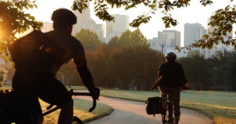 Two bicyclists wearing helmets riding bikes on the greenway with the Charlotte skyline in the distance