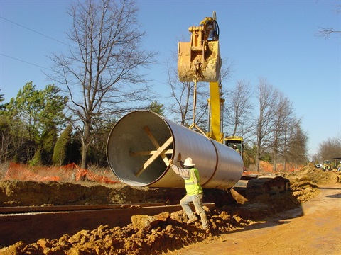 A backhoe lowering a very large pipe into a trench, guided by a working wearing a safety helmet and vest