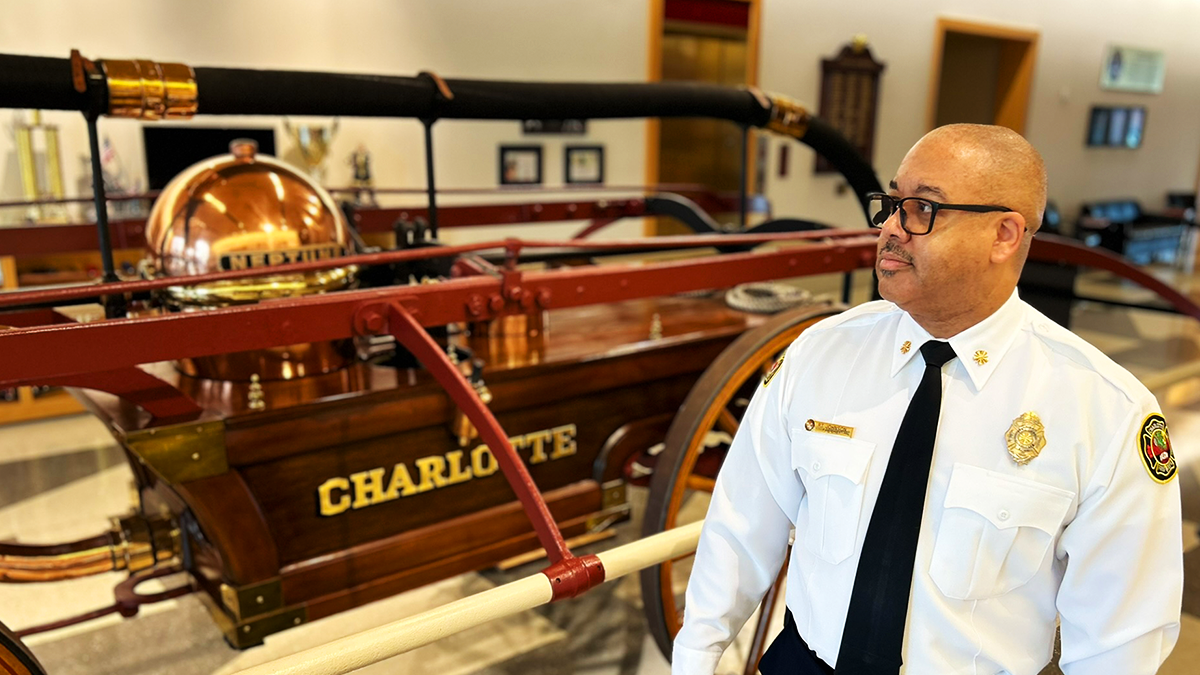 Charlotte Fire Chief Reginald T. Johnson stands in front of the historic Neptune hand-pumper at Charlotte Fire Headquarters, a lasting symbol of the legacy and resilience of Black firefighters in the city's history.