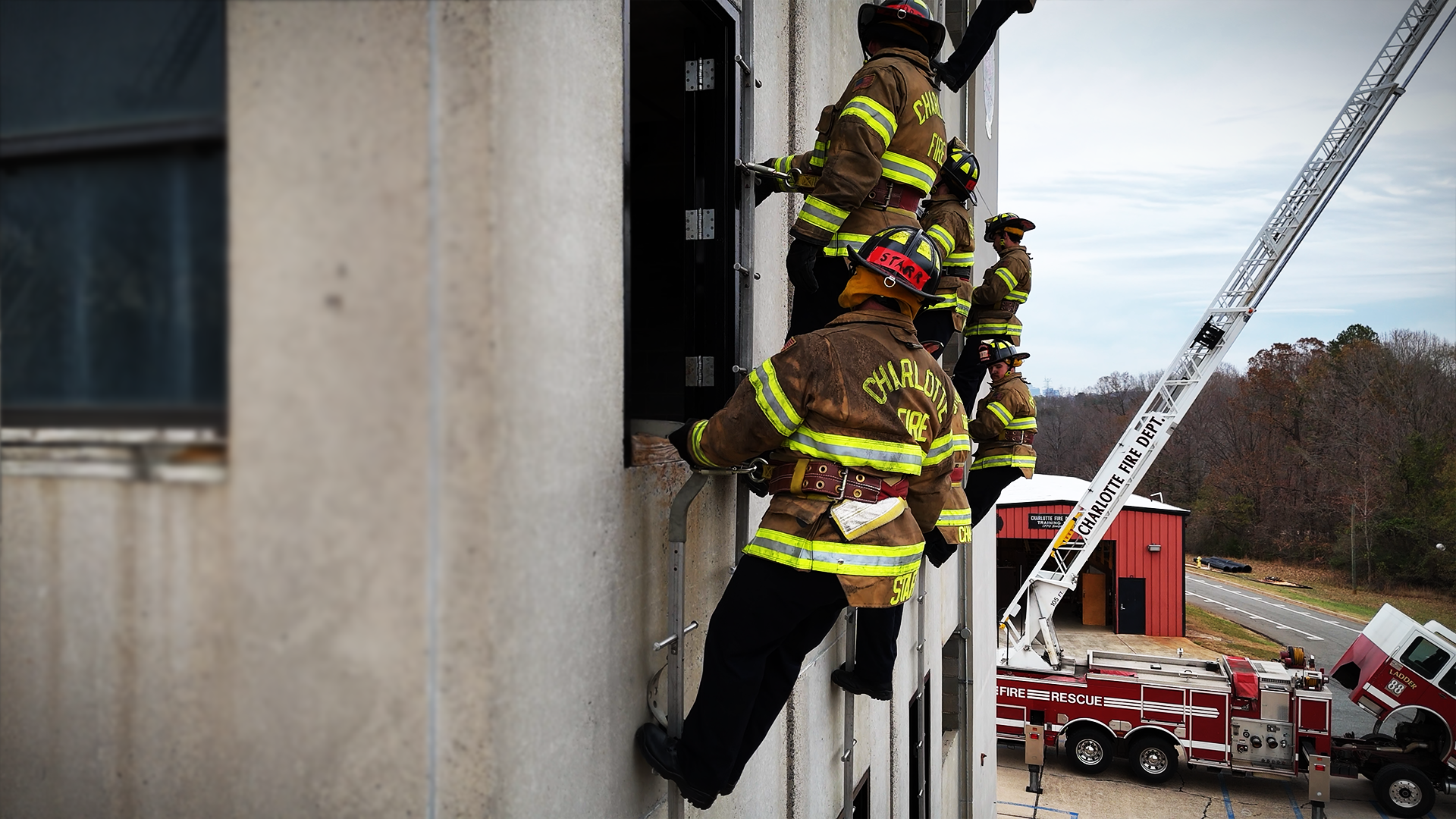 Fully geared recruits stage along the tower face, waiting for their turn as instructors coach each climber through the sequence