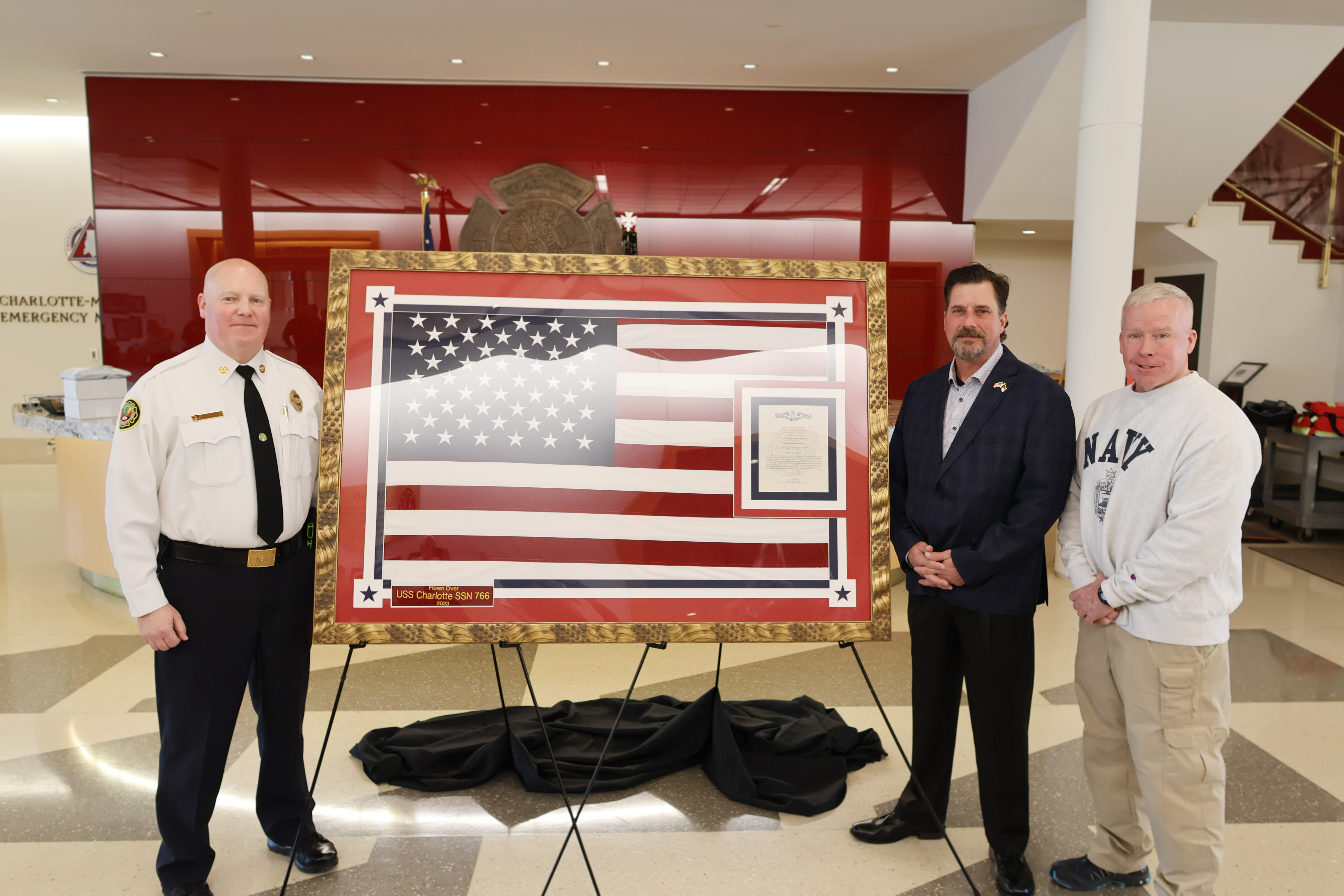 Charlotte Fire Superintendent Bill Suthard with retired Charlotte Fire Battalion Chiefs Shawn Royall and Rob Cannon stand with the framed U.S. Navy Ensign flown aboard the USS Charlotte during a dedication ceremony at Charlotte Fire Headquarters. 