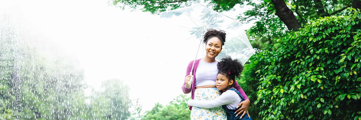 An adult and a child standing under a clear umbrella in the rain near green trees.