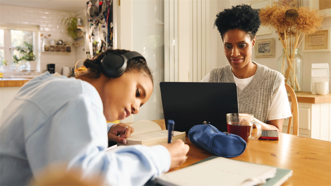 Mother at dinner table using laptop sitting beside daughter writing in notebook