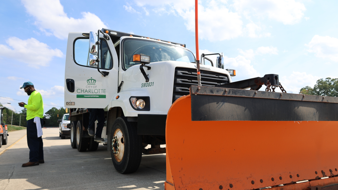 Snow plow during winter preparation training activity. A man stands beside the plow in bright colors while another person enters.