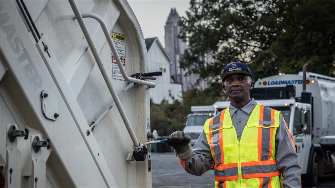 A sanitation worker poses next to a collection vehicle ahead of a morning shift. He wears a yellow safety vest and holds a lever on the vehicle.