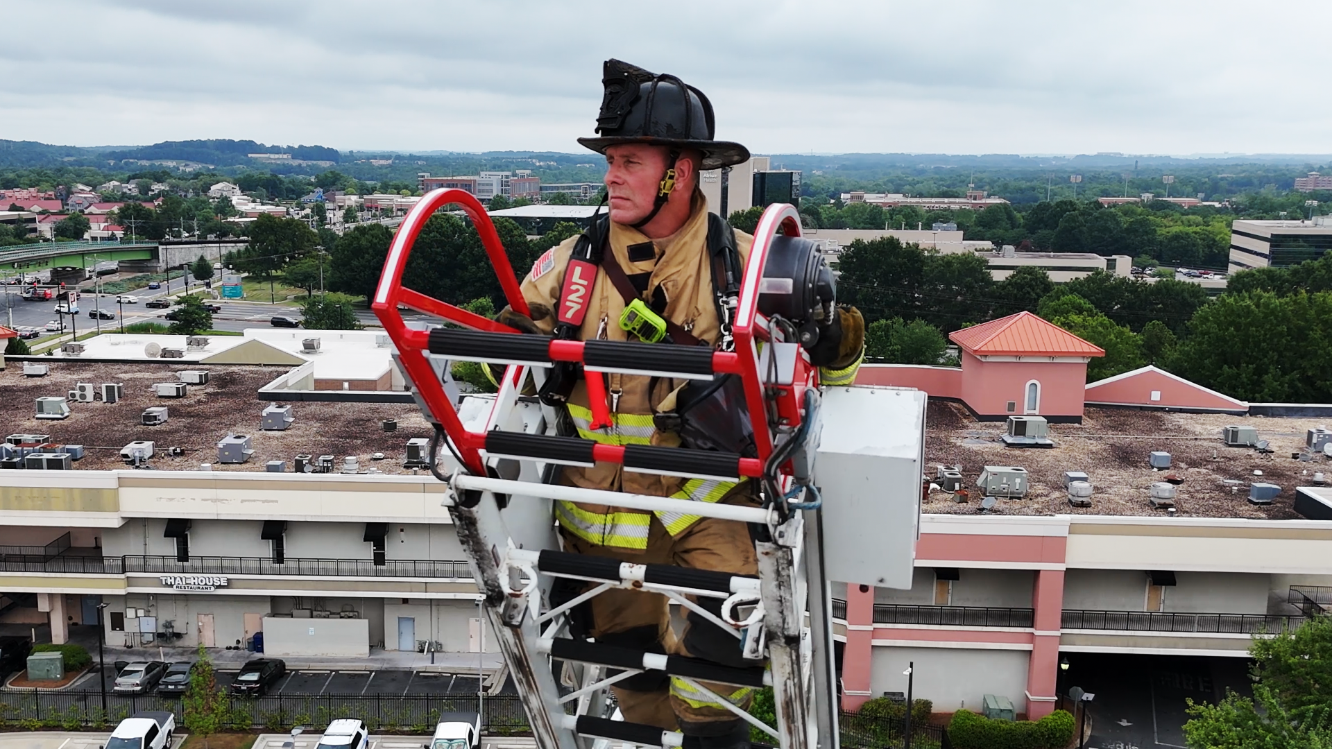 Wiatrowski in full firefighter gear on top of an elongated fire engine later. He is high above all the surrounding buildings and trees.