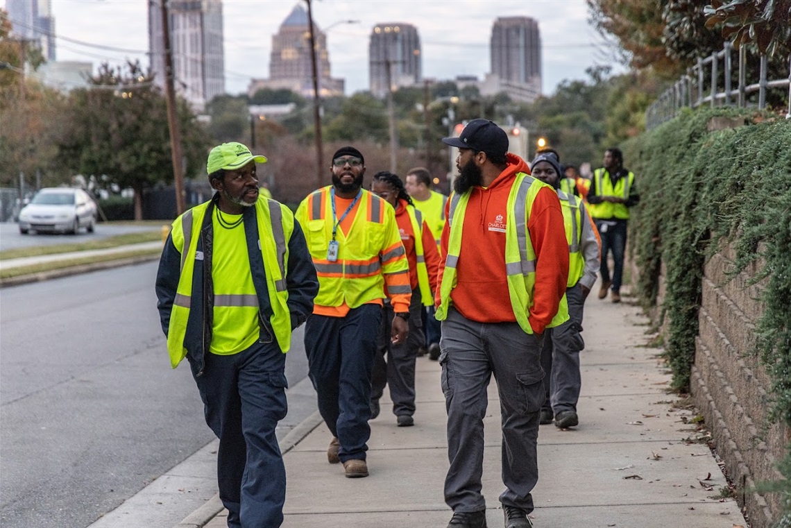 Several Solid Waste Services workers walk together down a sidewalk. The city skyline is behind them. They are all wearing bright yellow safety vests.