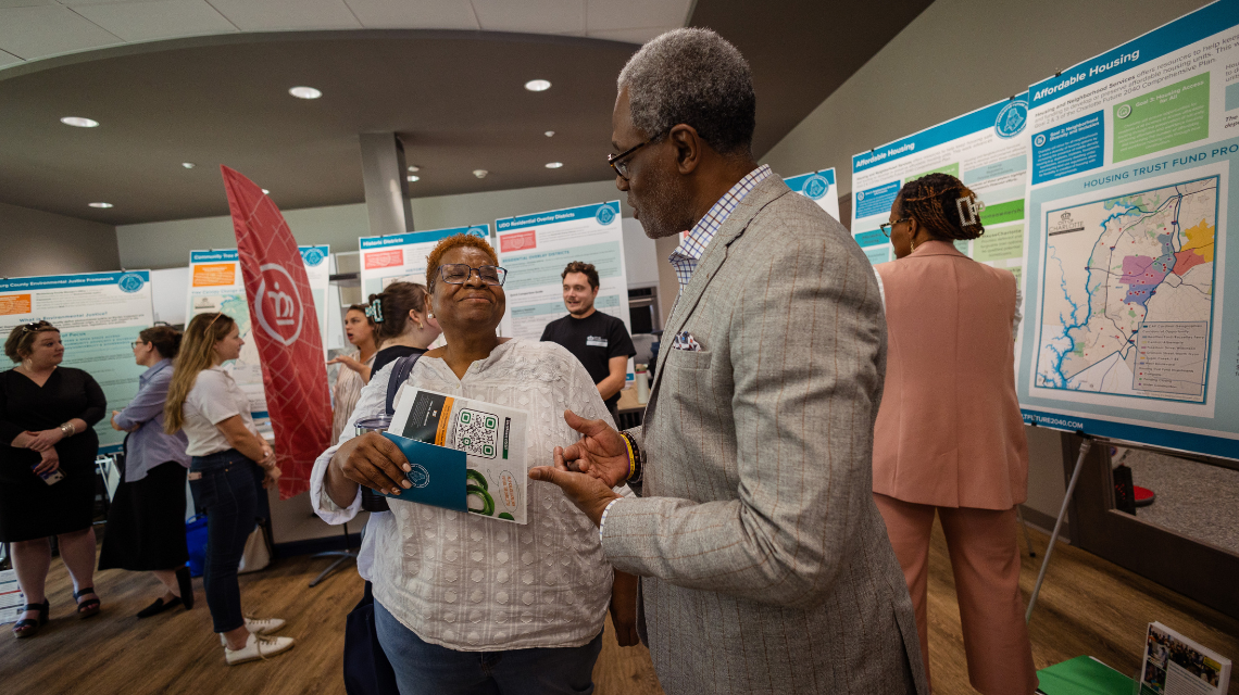 Two people discuss city planning during a community workshop. Maps and attendees surround them.