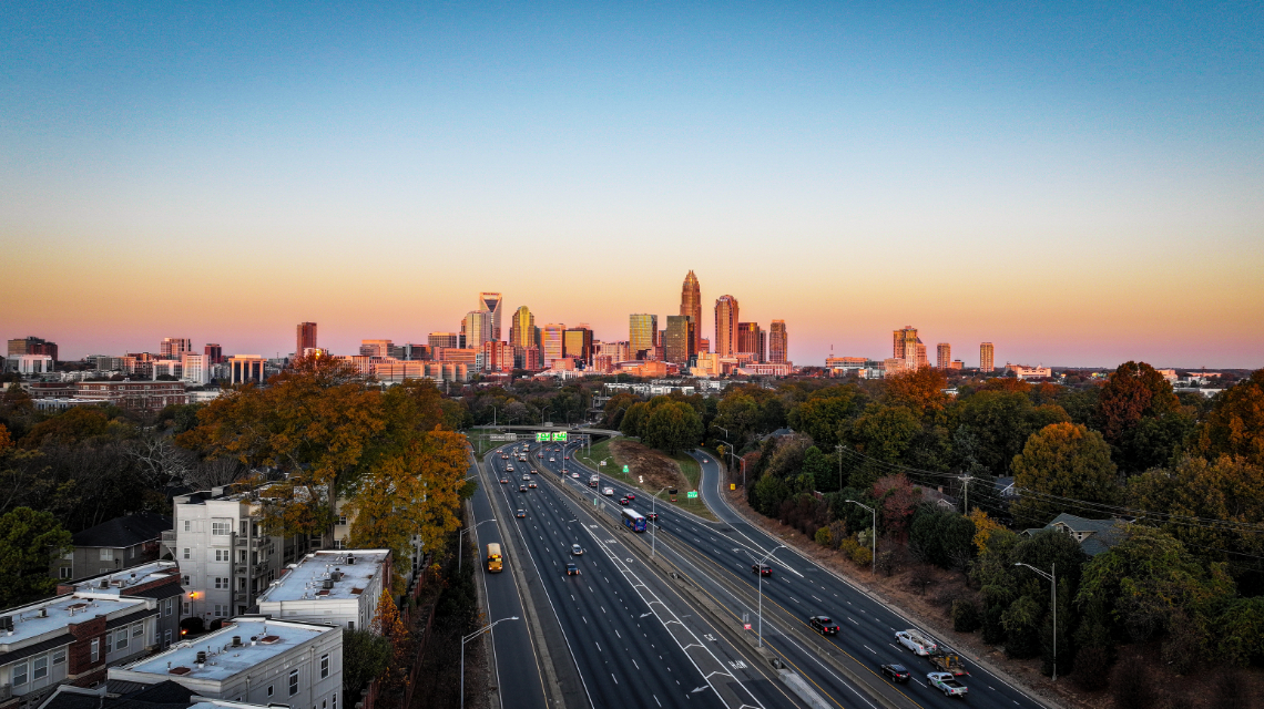 Charlotte skyline in the distance as the sun sets, creating pink and blue backdrop. In front of the city are roadways and buildings.