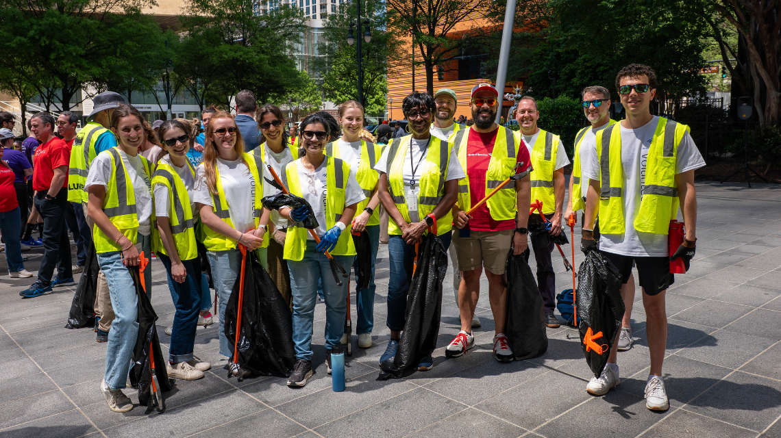 Volunteers in safety vests hold litter picking tools as they pose for a photo in Uptown Charlotte. A large crowd of volunteers is visible behind them.