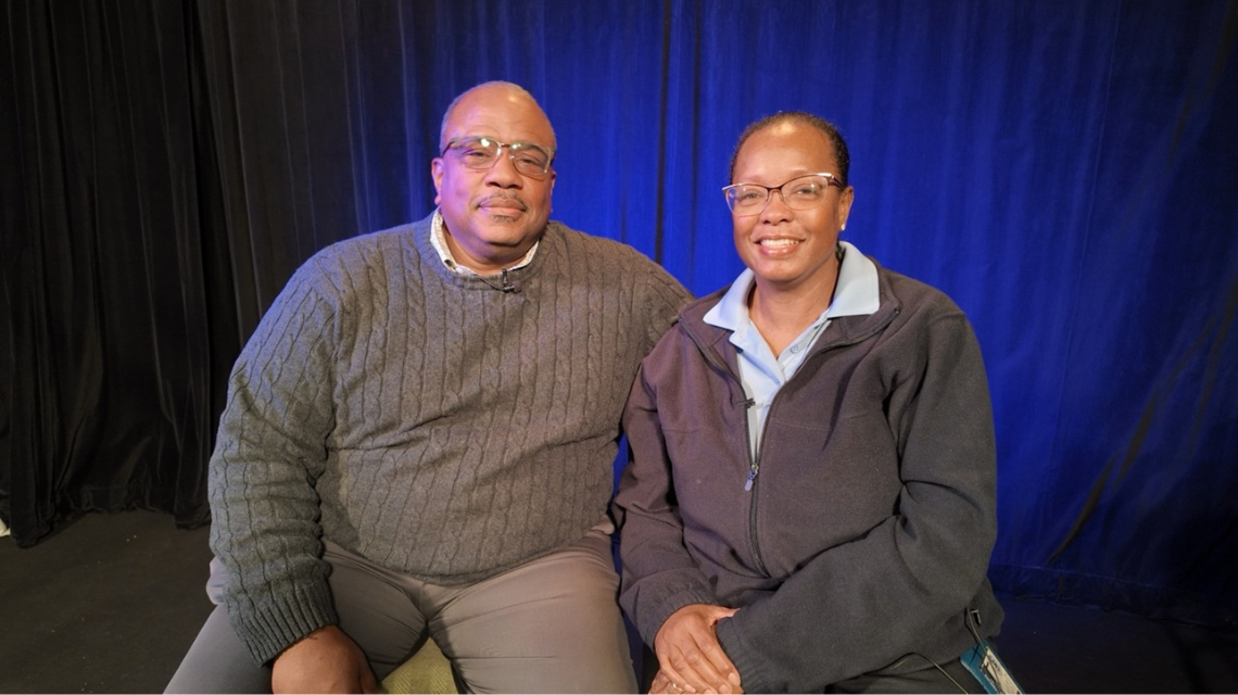 Robert and Monica Mahoney pose in a studio and smile at the camera.