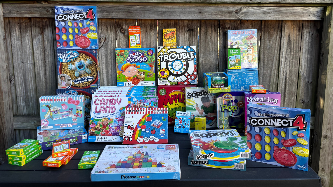 A variety of board games piled on a table in front of a wood fence.