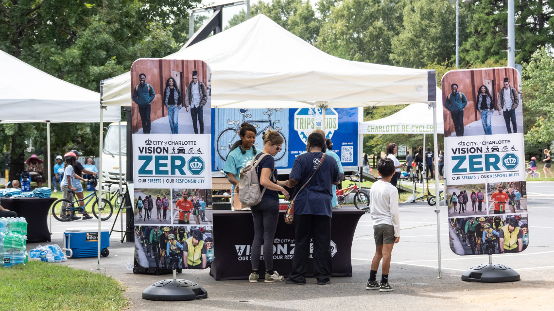 Attendees gather at a City of Charlotte Vision Zero booth during Mobility Safety Fest. Many people gather in the background with bikes and helmets.
