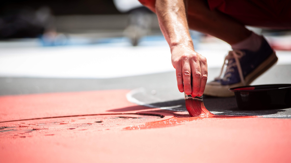 Close up of a person's hand painting a street mural with a brush. The paint is bright red.