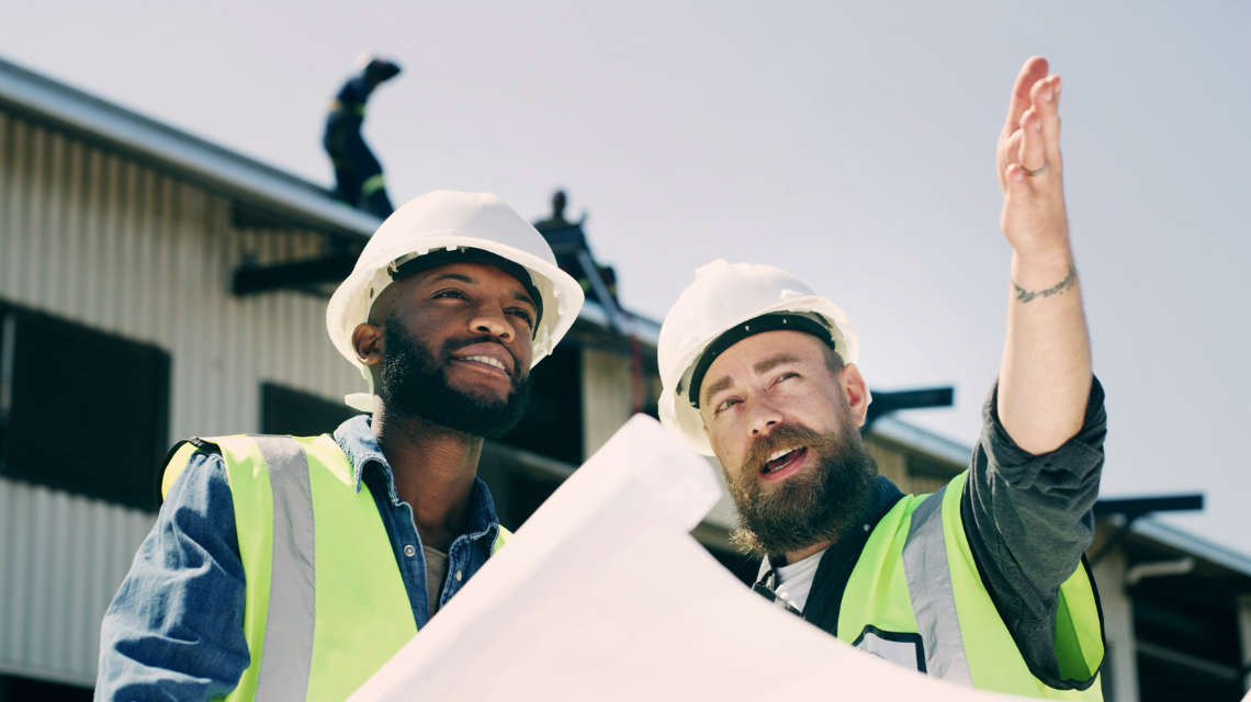 Two men in safety vests and hardhats review plans at a construction site. One man is instructing the other.