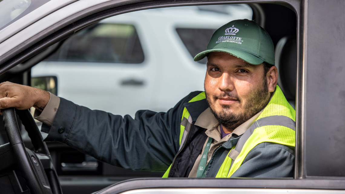 Solid Waste Services employee drives a truck while working. He has a Solid Waste hat and safety vest.