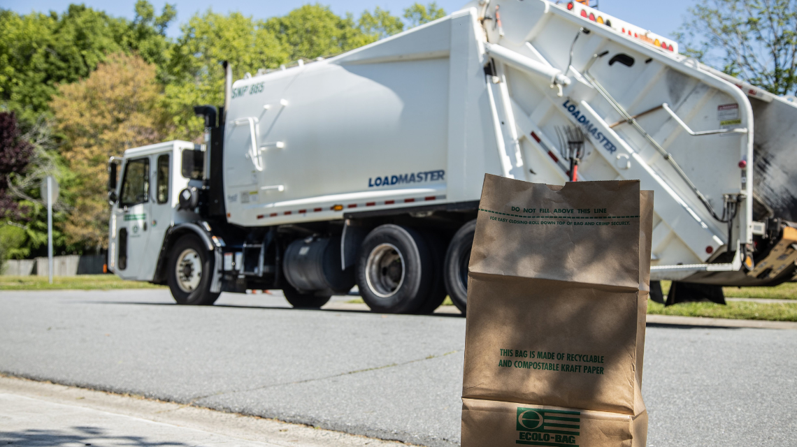 Brown yard waste bag sits curbside as a white collection truck passes on the street.