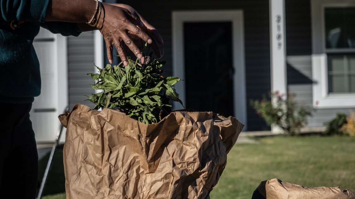 A woman's hands place leaves into a brown paper bag for yard water collection.