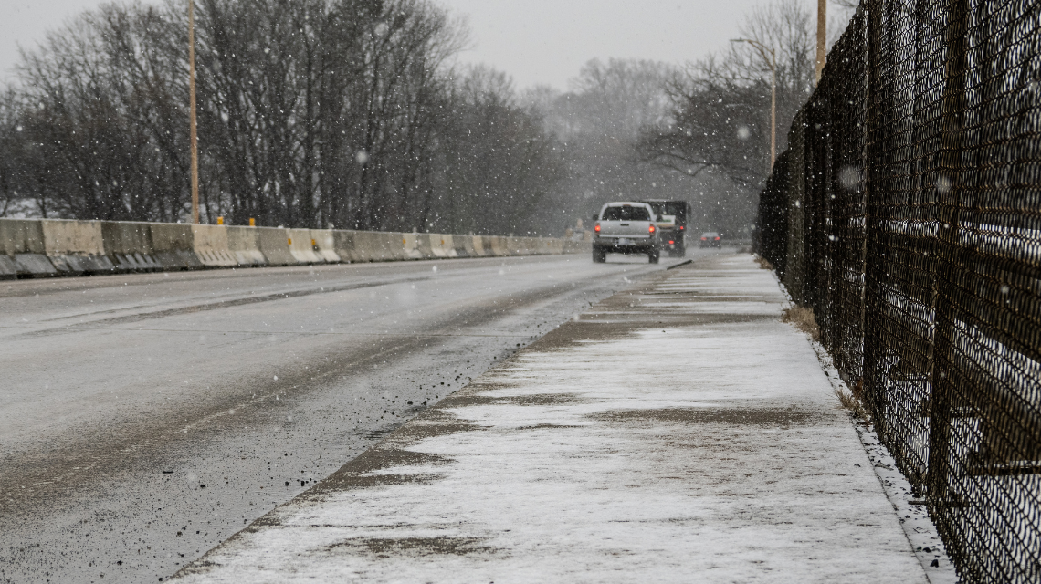 Snow falling on a Charlotte road and collecting on the sidewalk as vehicles drive away in the background.