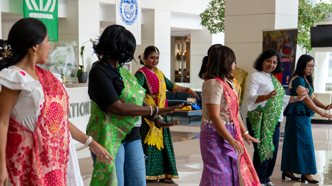 A diverse group of women in cultural attire dance together in the Government Center lobby during an event. Banners displaying the City of Charlotte and Mecklenburg County logos are visible behind them.