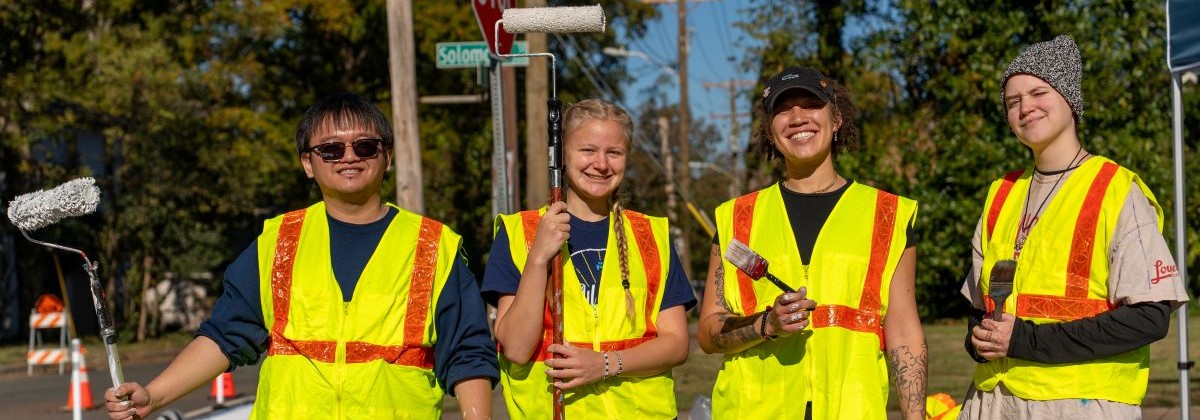 A group of men and women in safety vests posing with paint brushes and rollers standing in the street..