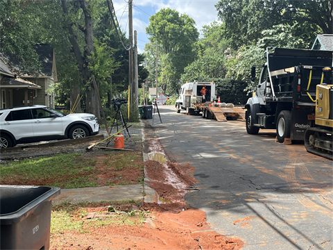 Residential street with utility work: Charlotte water trucks and equipment parked roadside, workers near a service vehicle, and muddy runoff along the curb under leafy trees due to a water main break.