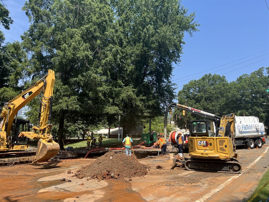Queens-Morehead roadwork crew repairs an underground utility line, with two excavators, a vacuum truck, hoses, and piles of dirt beneath tall trees on a sunny day.