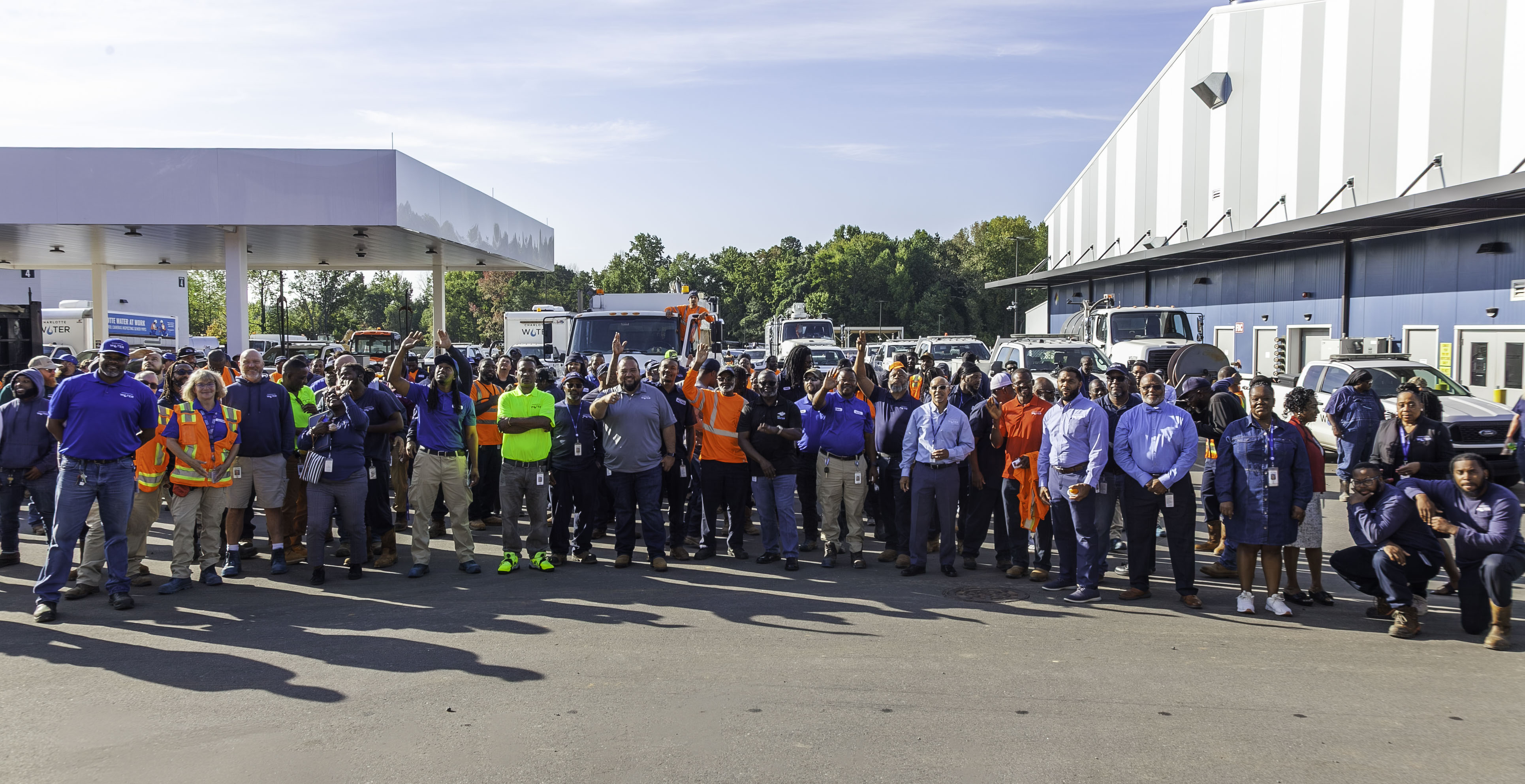A large group of Charlotte Water workers, many in safety vests and uniforms, stand together outside near trucks and a building on a sunny day.