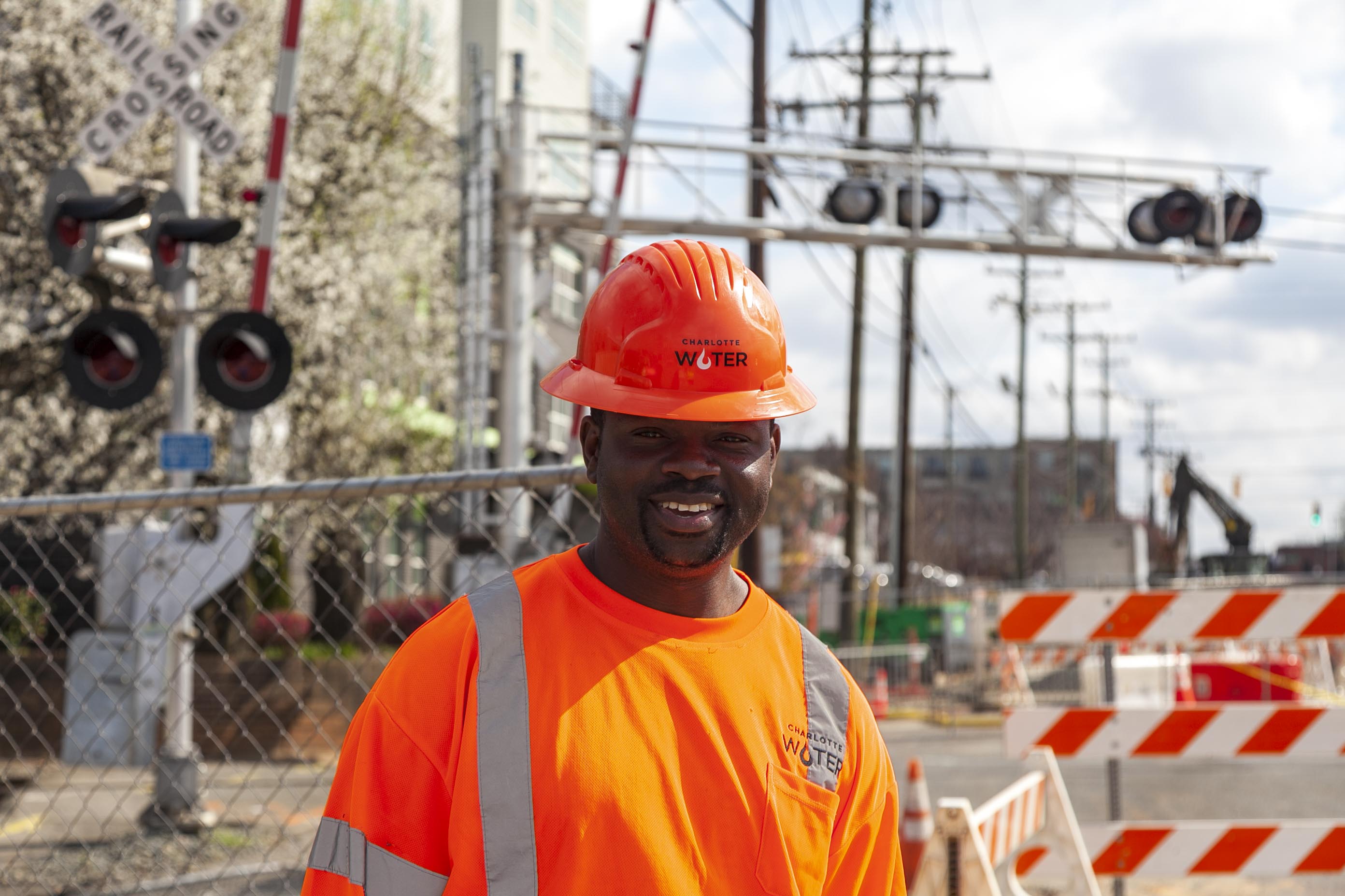 A Charlotte Water worker, smiling, wearing an orange safety vest and a hard hat, stands in front of a railroad crossing and traffic barriers at a work site.