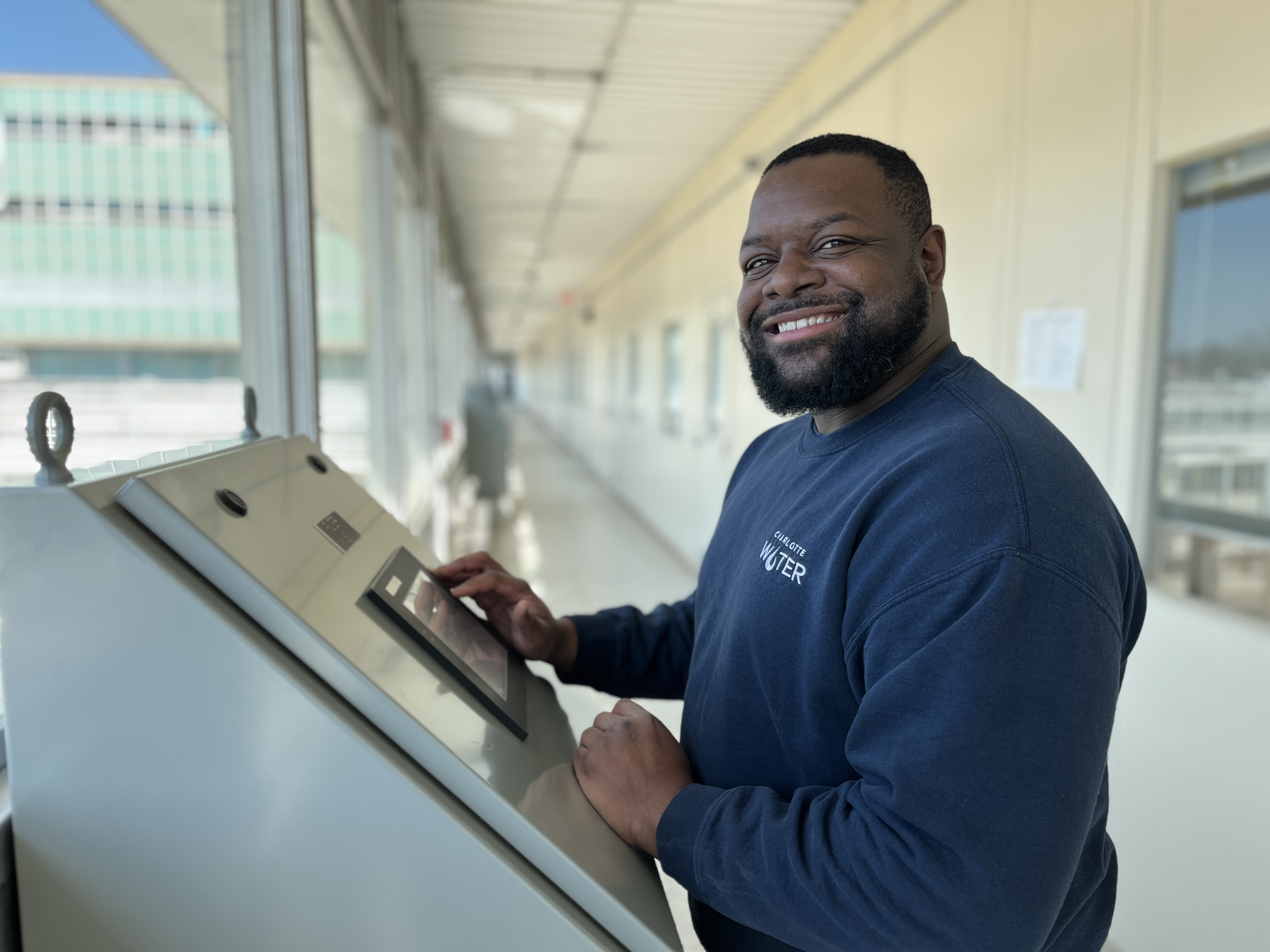 A Charlotte Water worker, smiling, wearing a blue Charlotte Water sweater, stands in front of machine at a water treatment plant.